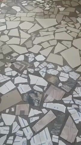 The video shows a close-up, top-down view of a floor tiled with broken pieces of ceramic and stone. The tiles are irregularly shaped and fitted together, creating a mosaic-like pattern. The colors vary, with a predominant use of white, beige, and gray, interspersed with darker, speckled pieces and some tiles with blue and white linear patterns. The grout is visible in the spaces between the broken pieces. The lighting suggests an indoor environment. No people or actions are visible in the frame.