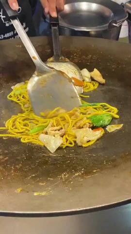 A food vendor is cooking noodles on a large, flat, circular griddle. They use two large metal spatulas to stir and toss the noodles, which are yellow and appear to be in a sauce. Mixed with the noodles are pieces of chicken, shredded cabbage, carrots, and green vegetables. The griddle itself is stained with oil and residue from previous cooking. The person cooking is wearing a dark-colored apron and can be seen lifting a lid and pouring liquid onto the griddle, presumably to add moisture and flavor to the dish.