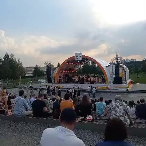 The video shows an outdoor jazz festival. A stage with a large arched canopy is set up at one end of a park. The canopy is white with orange accents and displays the word "JAZZ" in large letters. A band is performing on stage, with musicians visible behind their instruments. Several spotlights illuminate the stage. In front of the stage, a large crowd of people is seated on the ground, facing the performance. They appear to be enjoying the music. The park setting includes trees and a grassy area. The sky is cloudy, suggesting it might be late afternoon or early evening. The overall atmosphere seems relaxed and festive.