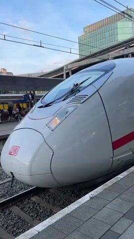 The video shows a white Intercity Express (ICE) train stopped at a platform. The train has a red stripe along its side and the number 8021 is painted on it. People are walking along the platform, some boarding the train. A digital sign displays information about the train's destination: Amsterdam Centraal, with a departure time of 19:38 on platform 14. The platform is tiled in gray and white. Above the platform is a modern canopy structure. In the background, there is a glass-walled building and overhead power lines. The sky is slightly overcast.