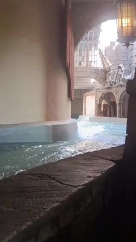This media file captures a scene from a water park, likely at Phantasialand in Brühl, Germany, given the distinctive theming. The setting appears to be an indoor attraction with a medieval or fantasy architectural style, characterized by stone arches and a castle-like structure in the background. A waterway, possibly a lazy river or a ride channel, flows through the scene, generating gentle waves.

The subject of the video is the flowing water and the environment it's situated in. While no distinct people are clearly visible or the focus, the presence of what appear to be structural elements and perhaps distant figures suggests this is an active amusement park area. The lighting suggests it could be daytime, with natural light filtering in from above, though the enclosed nature of the area makes it hard to determine the exact time of day or weather conditions.

Notable details include the rough-hewn stone textures of the architecture, the wooden elements near the left edge, and a decorative lantern hanging from the ceiling on the right. The water itself is a prominent element, its movement and texture contributing to the overall atmosphere. The scene evokes a sense of adventure and escapism, common to theme park experiences. There is no visible text within the media file.