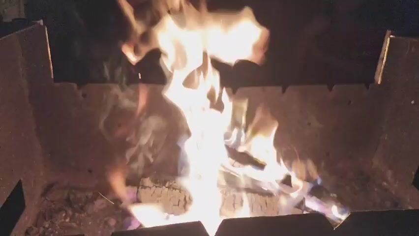This video shows a close-up, medium shot of a bonfire in a metal fire pit at night. The fire is burning brightly with flames of yellow, white, and hints of purple. Logs and embers are visible within the pit, and smoke is rising. The background is dark and out of focus, with some blurry shapes that might be people or objects. The camera is stationary.