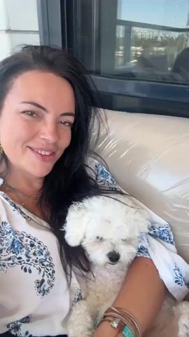 A woman with long, dark hair and wearing a white top with a blue floral pattern is holding a small white dog. She is smiling at the camera. In some frames, a man with short blonde hair and a beard, wearing a light green t-shirt, appears next to her, making a surprised expression. The background suggests an outdoor setting with a building facade and a window.