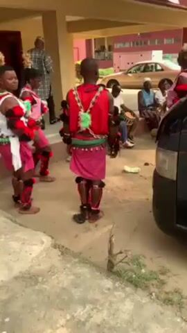 The video shows a group of dancers performing outside a building. The dancers are wearing traditional outfits featuring red, pink, and white colors with fringe and ornaments. They are barefoot and appear to be moving to the beat of a drum played by a seated musician in the background. Several other people are watching the performance.  A dark-colored van is parked nearby. The setting seems to be a paved outdoor area near a building with a covered walkway.