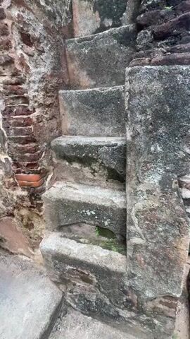 The camera pans up a flight of exterior stone stairs affixed to a brick wall, revealing an ancient structure. The stone steps show signs of age, with visible moss and staining. The brick walls are also worn, with some bricks missing and evidence of repair or patching. The sky is visible in the upper portion of the frame, suggesting the structure is tall.