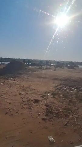 The video shows a sunny day at a sandy beach or riverbank. Several people are gathered by the water's edge. There are a few vehicles parked in the background. The ground is mostly dirt and sand with some vegetation. The sun is bright in the clear blue sky. There is music playing in the background.