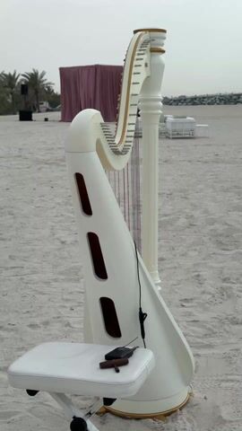 The video shows a white harp and matching stool set on a sandy beach. The harp is positioned to the right of the stool. The stool has a small, dark object on top, possibly a tuner or microphone. In the background, there is a pink fabric structure, palm trees, and some white furniture. The sky is overcast. The scene suggests a setup for a musical performance or event on the beach. The camera is static, capturing the scene from a slightly elevated angle.