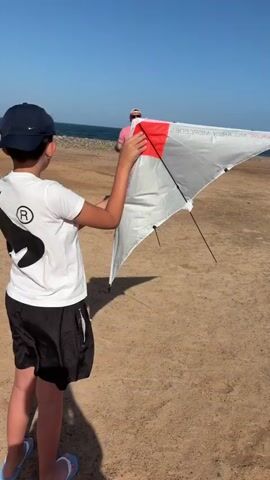 A young boy in a white t-shirt, black shorts, and a navy baseball cap stands on a sandy beach, holding a white and red delta-shaped kite. An adult male in a pink shirt, khaki shorts, and sunglasses stands several feet behind the boy. The ocean is visible in the background under a clear blue sky.