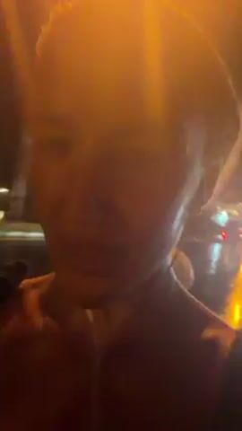 A person is standing outside at night. The light is poor. The person appears to be fair-skinned, female, with light hair pulled back into a ponytail. They are wearing earphones and a black tank top. There is a bright light in the background. The sound of thunder and rain can be heard.