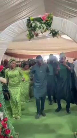 A group of people dance at a festive indoor event under a white, flower-adorned archway. Most are dressed in shades of green, including three women at the front wearing elaborate lace outfits and headwraps. Two men in the center of the group wear modern tunics and dark sunglasses, while others behind them are dressed in traditional Yoruba attire including "fila" caps. The setting looks like a large tent or hall, filled with guests seated at tables in the background. The overall atmosphere is lively and celebratory.