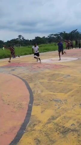 A group of young men are playing basketball on an outdoor court. The court surface is a mix of yellow and orange concrete with red boundaries. In the background, there is a basketball hoop with a torn net and some trees and buildings. The sky is overcast with dark clouds. The players are mostly shirtless, wearing shorts, and are actively engaged in the game, dribbling, passing, and shooting the basketball.