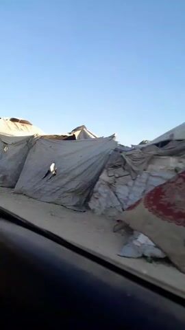 This video captures a scene in Deir el-Balah, Palestinian Territories, likely a displacement camp. The setting is characterized by a series of makeshift tents and shelters constructed from canvas and other tarpaulin-like materials, erected on sandy ground. The sky is a clear, bright blue, suggesting a sunny day with no clouds.

Several individuals are visible within the scene. At one point, a young girl in a blue and yellow patterned dress walks along a path between the tents. Later, a woman in a headscarf is seen sitting on the ground next to a blue water container and a red bucket, with a person in a wheelchair beside her. Another woman, also wearing a headscarf, carries a large blue container and other items as she walks through the camp. The tents themselves show signs of wear and tear, with some having visible rips or openings. There are also objects such as poles supporting the shelters, bags of what appears to be sand or grain, and laundry hanging outside one of the tents. The overall impression is one of a temporary and potentially challenging living situation for the inhabitants.