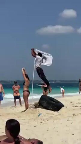 The video shows a beach scene on a sunny day. A man in a white shirt and black pants executes a backflip on the sand. Surrounding him are other people: a woman in a black bikini raises her arms, a man in blue shorts holds a phone, and another man in patterned shorts also has a phone. A woman in a black bathing suit stands nearby. In the foreground, a person reclines on the sand. A white flag with a logo waves in the background, next to a tire. The turquoise water of the ocean stretches to the horizon, dotted with boats.