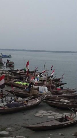 The video shows a riverbank cluttered with wooden fishing boats, each adorned with a black, red, and white flag. Some boats are piled with nets and other fishing gear. People are moving about on the sandy shore, some standing near the boats, others walking along the bank. In the distance, a larger boat or ferry is visible on the water. The overall scene suggests a fishing community going about their daily activities. The sky is overcast, giving the scene a somewhat muted appearance.