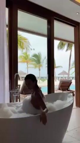 A woman with dark hair is sitting in a white, freestanding bathtub filled with bubbles. Her back is towards the camera. The bathroom has large sliding glass doors that open to an outdoor area with palm trees, lounge chairs, and what appears to be a pool.  The sky is overcast.  A modern faucet is visible next to the tub, and white curtains hang to the side of the doors. The scene suggests a luxurious vacation setting.