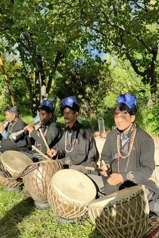 The video shows a group of four young men sitting in a grassy, outdoor area with trees overhead. They are all wearing traditional clothing, including dark robes and blue hats. The men are playing musical instruments. The two on the left are playing flutes or reed instruments. The two on the right are playing drums with drumsticks. The men appear to be playing in unison. The lighting suggests it is daytime.