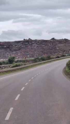 The video shows a road curving to the right, with a rocky hillside on the left. There are guardrails and vegetation along the side of the road. Yellow curve warning signs are visible on the hillside. The sky is cloudy. A white bus appears in the distance on the right side of the road.