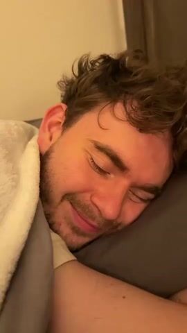 A man is lying in bed, sleeping. His eyes are closed, and he is smiling. Someone's finger is poking his forehead, causing him to react with a wince and then a laugh. He is covered by a blanket.