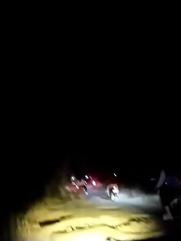 A vehicle's headlights illuminate a dirt road at night, revealing a convoy of motorcycles. The first few motorcycles are visible, followed by others further down the road. Some of the motorcycles appear to have riders, while others may be empty. The road is uneven, with visible patches and debris. The scene suggests a group traveling together in a rural or unpaved area under the cover of darkness.