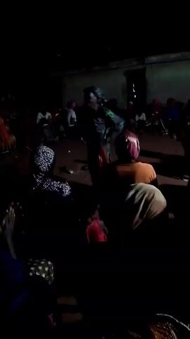 The video shows a dimly lit gathering of people, seemingly outdoors, perhaps in a courtyard or open space.  Most individuals are seated, facing a person standing in the center who appears to be addressing the group.  The speaker is wearing a patterned garment and a head covering.  The other people in the shot are mostly in dark clothing, with some wearing head coverings as well.  The lighting is poor, making it difficult to discern details, but the building's construction seems basic.  There are some scattered objects on the ground. Some people in the crowd are clapping their hands.