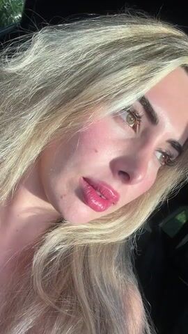 A close-up, low-angle shot captures a young woman with blonde hair and full lips. She is looking down and to the right, her eyes partially closed. The sunlight catches her face, highlighting her features. The background is dark, with hints of green foliage visible on the left. Her skin appears smooth with a slight pinkish hue on her cheeks. She is wearing glossy pink lipstick.