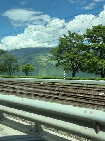 This is a video captured from a moving vehicle, likely a car or train, looking out towards a scenic landscape. The foreground is dominated by train tracks and a metal guardrail. In the middle ground, a serene lake reflects the bright blue sky, which is dotted with fluffy white clouds. Lush green trees line the edge of the lake, and rolling hills covered in verdant fields rise behind them. In the distance, mountains create a dramatic backdrop, their peaks partially obscured by the haze of the day.

The time of day appears to be midday, given the bright sunlight and clear skies. The weather is pleasant and sunny, suggesting a warm day. The presence of train tracks and overhead power lines indicates a location with active transportation infrastructure, and the specific mention of Zell am See, Austria, places this scene in a European alpine setting known for its natural beauty.

There is no discernible activity or event taking place beyond the movement of the vehicle capturing the video. The overall emotion evoked by the scene is one of tranquility and appreciation for nature's grandeur.

Visible text includes "98 9" on a small white sign next to a tree on the left side of the frame, and some faint markings on a concrete pillar on the right side of the frame.