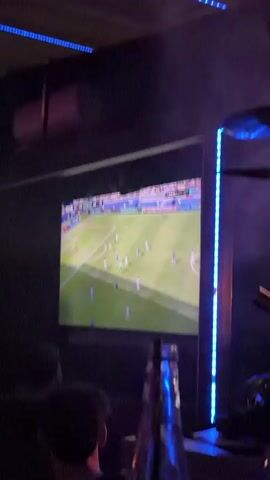 This is a video of a television screen displaying a soccer match. The game is being played on a green field with players in white and blue uniforms. There is a stadium in the background with spectators. The score on the screen indicates 0-0. In the foreground, there are people watching the television. The room is dark and illuminated by the television screen and blue LED lights.