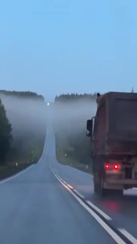 The video shows a view from a vehicle driving on a wet asphalt road through a foggy forest. The road curves to the right and then straightens as it goes uphill into the fog. There are trees on both sides of the road. In the foreground on the right, the rear of a large truck is visible, with its red taillights illuminated. The sky is a pale blue and the overall lighting suggests it is early morning or late evening.