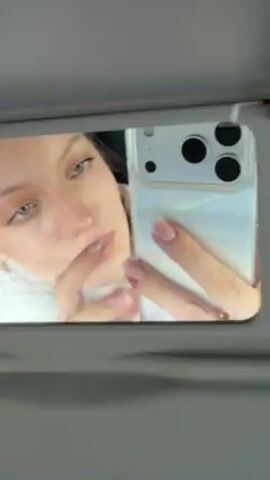A young woman takes a selfie in a car mirror. She is fair-skinned with light eyes and pink lips. She wears small earrings and a ring. The phone is encased in a clear, glittery case. Light conditions change from bright to dim, altering the shadows on her face. The background is primarily dark, suggesting an interior car setting. Her hand is visible holding the phone and resting against her cheek at times.