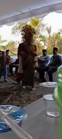 The video captures a wedding ceremony taking place outdoors under a white tent. A bride, adorned in a traditional, ornate gold headdress and a richly decorated burgundy and gold outfit, walks forward. She is accompanied by a groom in a matching burgundy suit and traditional beaded necklaces. Both are holding white floral garlands with red accents. Several men, some in traditional attire and some in casual clothing, are seated and standing around the ceremony area, observing the procession. The ground appears to be dirt and gravel. In the foreground, partially visible, are a clear plastic cup with a drink and a blue object, possibly another cup or container. The overall atmosphere suggests a festive cultural event.
