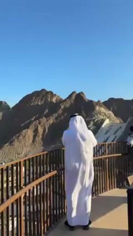 A man in traditional white Middle Eastern clothing stands on a platform overlooking a valley with a mountain range in the background. The platform has a brown metal railing. The man is standing with his back to the camera and appears to be looking at a sign or display on the platform. The valley below contains white buildings with trees and shrubbery. A winding road can be seen going up the side of one of the mountains. The sky is clear and blue.