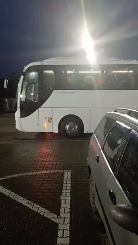 A white bus and a silver station wagon are parked side-by-side in a parking lot at night. The bus is on the left, and the station wagon is on the right. The bus has its headlights on, and there are lights on inside the bus, illuminating the windows. People can be seen sitting inside the bus. The station wagon's windows are wet, suggesting it may have rained recently. The parking lot is paved with dark bricks, and there are white parking lines visible. A bright light source, possibly a streetlight or floodlight, is shining down from above, creating a glare in the sky.