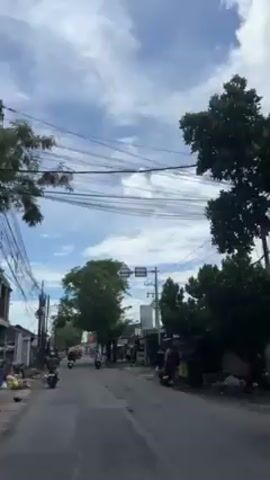 The video shows a street scene viewed from a moving vehicle, likely a motorcycle. The road is paved and relatively clear, with other motorcycles visible moving in the same direction. Buildings, some with awnings and signs, line the sides of the street. Trees with full green foliage are interspersed along the road, partially obscuring the buildings. Overhead, a network of electrical wires crisscrosses the sky, which is mostly blue with scattered white clouds. The overall impression is of a casual daytime street scene in a residential or commercial area.