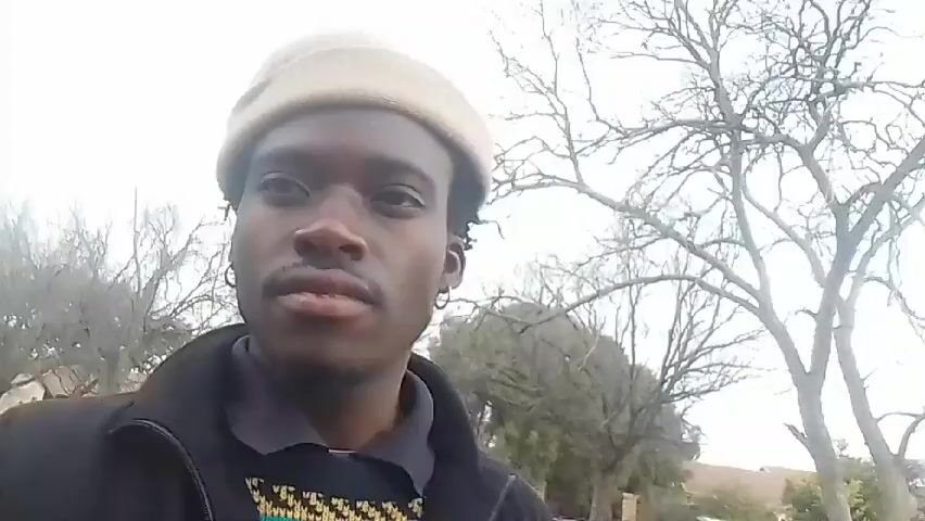 The video features a young Black man filming himself outdoors in a selfie style in what appears to be a residential area of Klerksdorp, South Africa. He wears a light-colored beanie, a black jacket over a patterned sweater (featuring green, red, yellow, and white geometric designs), black pants, and black sneakers. He also has silver hoop earrings.

The background shows houses, fences, and various trees, some with bare branches and others with green foliage, under an overcast sky. The ground is a mix of dirt and possibly paved areas.

The man initially walks forward, looking at the camera with a neutral expression. The camera then rotates to show his full outfit and the surrounding environment, including a brief upward shot of the cloudy sky. He strikes a pose, standing with arms extended and then makes a short, rhythmic movement or dance step. The camera rotates back for a close-up, and he concludes the video by smiling broadly at the camera.

Visible text on his clothing includes "NYC USA" and "KING" on his black pants, and a word resembling "heretel" along with a small graphic on his black jacket/sweater.