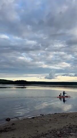 Two figures are visible in a red kayak on a calm body of water. The person in the back of the kayak appears to be paddling. The water is reflecting the cloudy sky. The shoreline in the foreground is sandy with some pebbles. A dark treeline is visible in the distance across the water. The sky is filled with a mix of dark and lighter clouds.