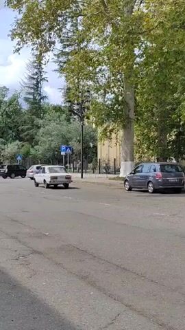 The video shows a street scene with cars driving. A white sedan is turning to the left, and a red hatchback is driving forward. There are trees and buildings in the background. The road appears to be asphalt.