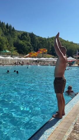 A man stands at the edge of a swimming pool, looking up and extending his arms above his head. He then leaps into the pool, creating a large splash. The pool is surrounded by lounge chairs and umbrellas, with a water slide visible in the background. Trees and a clear blue sky complete the scene.