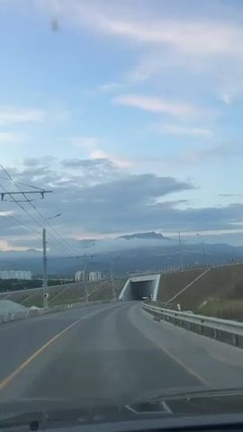 A vehicle travels down a highway, approaching a tunnel. The tunnel entrance is visible, and through the tunnel, a city can be seen. Mountains rise in the distance, partially obscured by clouds. The sky is a soft blue with scattered clouds. The highway is paved, with yellow and white lines marking the lanes. Guardrails line the sides of the highway. Overhead power lines are visible to the left. The lighting suggests it is either early morning or late evening. The video is filmed from inside the vehicle.