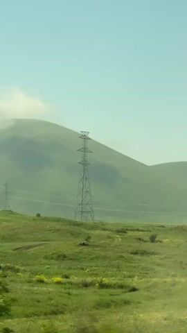 The video shows a wide, green, rolling landscape under a clear blue sky. In the foreground, there is a blur of green foliage, suggesting movement, possibly from a vehicle. In the middle ground, a tall, metal electrical pylon stands prominently in a grassy field dotted with yellow wildflowers. Another, similar pylon is visible further in the distance to the left. Gentle, green hills form the background, with a hint of haze obscuring the furthest ones. The overall impression is of a rural or natural environment with elements of infrastructure.