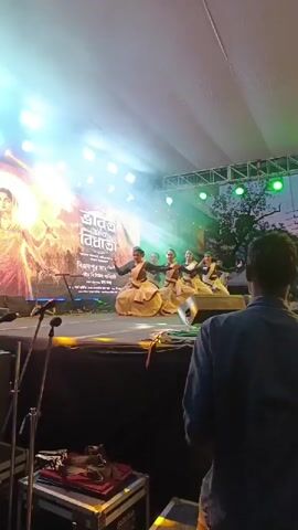 A group of six women in traditional Indian attire are performing a dance on a brightly lit outdoor stage. They are dressed in white and cream colored sarees with gold trim. The stage is set up with a large backdrop that displays an image of a woman in similar attire with fiery motifs and text in a non-English script. Several spotlights illuminate the performers, casting colorful light onto the stage. A few microphone stands are visible in the foreground, along with audio equipment. A person's back is visible in the lower right corner of the frame, facing the stage, suggesting an audience member or technician. The overall environment appears to be an outdoor event or festival.