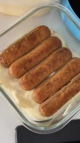 A close-up, overhead shot shows ladyfingers arranged in a clear glass dish. A white spatula is used to spread a creamy, pale yellow substance over the ladyfingers. The dish is filled with a layer of the same creamy substance at the bottom. The background is blurred but shows a white surface and part of a blue container. The action is focused on the preparation of a dessert.
