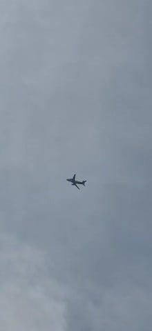 A low-angle shot shows a commercial airplane flying against a cloudy sky. The plane is silhouetted against the clouds and appears to be a passenger jet. A white contrail is visible behind the plane, indicating it has recently flown through the area. The plane is moving from left to right across the frame.