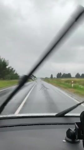 The video shows a view from inside a car driving on a wet road under a cloudy sky. The windshield wipers are intermittently clearing the windshield. The road is empty except for the car the footage is being filmed from. The surrounding landscape consists of fields and trees.