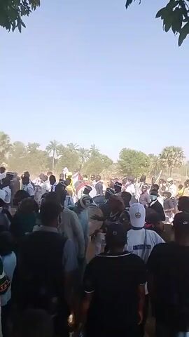 The video shows a crowd of people gathered in an outdoor area under a blue sky. There are trees and some visible buildings in the background. Most of the people are wearing dark clothing, some wear hats or caps. A few individuals wear white shirts or shirts with text on them. A band with drums is present in the crowd. People are dancing. There appears to be some dust in the air. Some people carry flags.