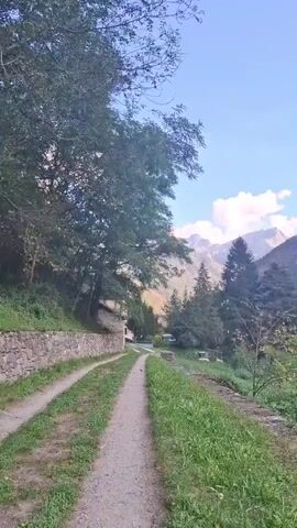The video pans across a serene landscape featuring a dirt path bordered by lush green grass and trees. In the background, majestic mountains rise under a clear blue sky with scattered white clouds. Several houses are visible, nestled among the trees and on the slopes of the mountains, suggesting a rural or small-town setting. The overall atmosphere is peaceful and idyllic, showcasing the natural beauty of a mountainous region.