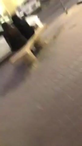 The video shows a street at night. There are cars parked along the street, and buildings are visible in the background. The street is paved with bricks. The camera is shaky and moves around. The lighting is dim, with streetlights providing illumination.