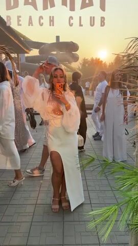 A woman in a white dress with a high slit and sheer sleeves is taking a selfie with her phone. She is wearing a shell-adorned top and a gold necklace. The background shows a beach club with water slides and palm trees. The sun is setting, casting a warm glow over the scene. Several other people, dressed in white, are visible in the background. One man is walking towards the left, and a woman in a white jumpsuit is standing on the right. The overall atmosphere appears to be festive and relaxed.