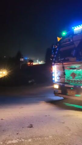 A vehicle, likely a truck with a tanker trailer, is shown at night. The truck is illuminated with various colorful lights, including blue, green, and white, which reflect on the wet road surface. The road appears to be a highway with lane markings visible. In the background, there are distant lights, suggesting a populated area or city lights. The overall scene depicts a vehicle on a road in low-light conditions, with evidence of recent precipitation.
