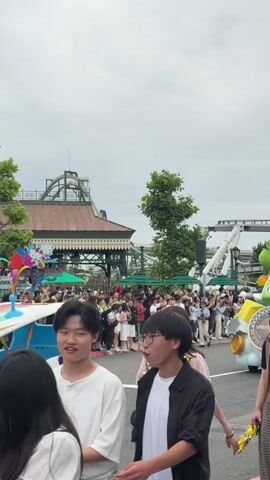A daytime scene captures a parade at what appears to be an amusement park, likely Universal Studios Japan, given the "UNIVERSAL" logo on the floats and the provided location context. The sky is overcast.

In the foreground, several spectators stand watching the parade. Two young men are partially visible on the left, and two young women are prominent on the right. One woman wears a light pink short-sleeved top and a yellow Pikachu necklace, while the other wears a black short-sleeved top, a plaid skirt, and yellow Pikachu wristbands.

Behind these spectators, a crowd lines the street, observing the procession. Two colorful parade floats are passing by. The first float features a large figure of Yoshi from the Mario franchise, seated in a kart designed to look like it's on clouds, adorned with gold coins and "UNIVERSAL" logos. A second similar float with Princess Peach is partially visible behind the Yoshi float. In the background, roller coaster tracks and other amusement park structures are visible, along with green trees and park buildings.