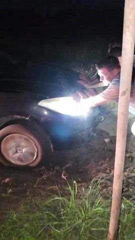 In a dimly lit, outdoor environment, possibly a dirt road or field at night, a dark-colored car is stuck in mud, its front right tire partially submerged. Two men are actively engaged in trying to free the vehicle. One man, wearing a grey t-shirt and light-colored pants, is positioned at the front of the car, leaning forward and appearing to push or pull on the headlight assembly. His face is illuminated by the car's bright headlight. Another man, wearing a white t-shirt, is visible behind him, also leaning in and possibly assisting. A third individual's head is visible near the driver's side of the car, suggesting they might be the driver or another helper. The ground around the car is muddy and uneven, with some grass and vegetation visible in the foreground and background. The overall impression is a group of people attempting a manual rescue of a stranded vehicle.